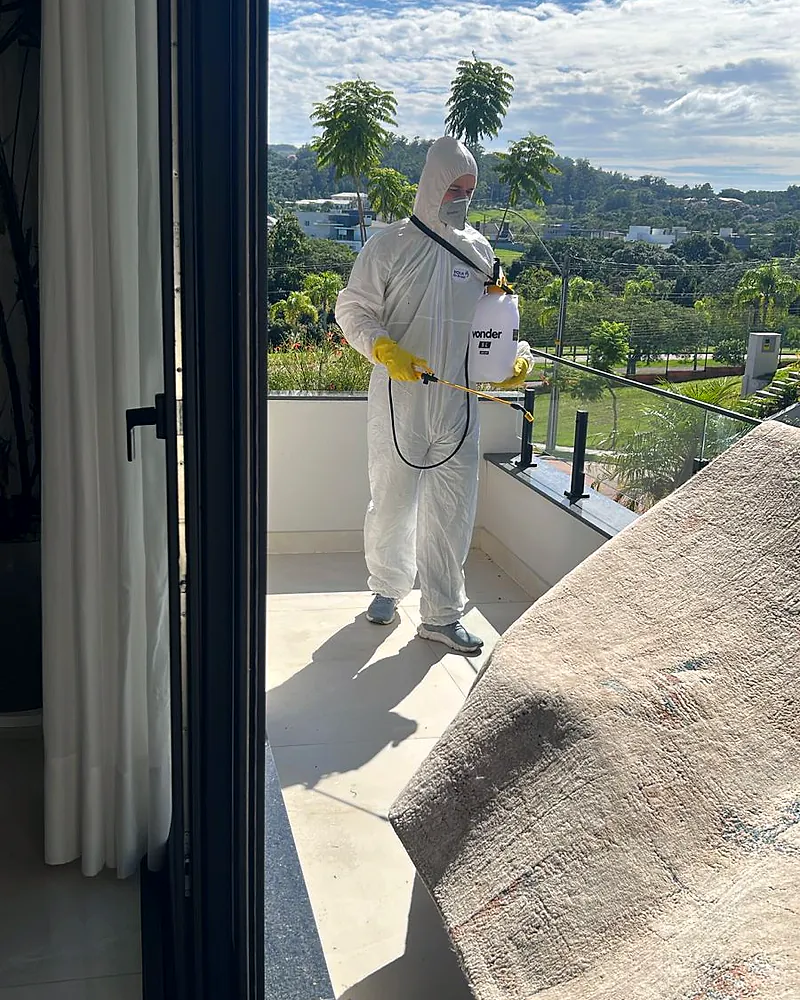 Técnico de macacão branco e pulverizador costal aplica produto na varanda de um apartamento, visto de dentro através da porta de correr aberta.