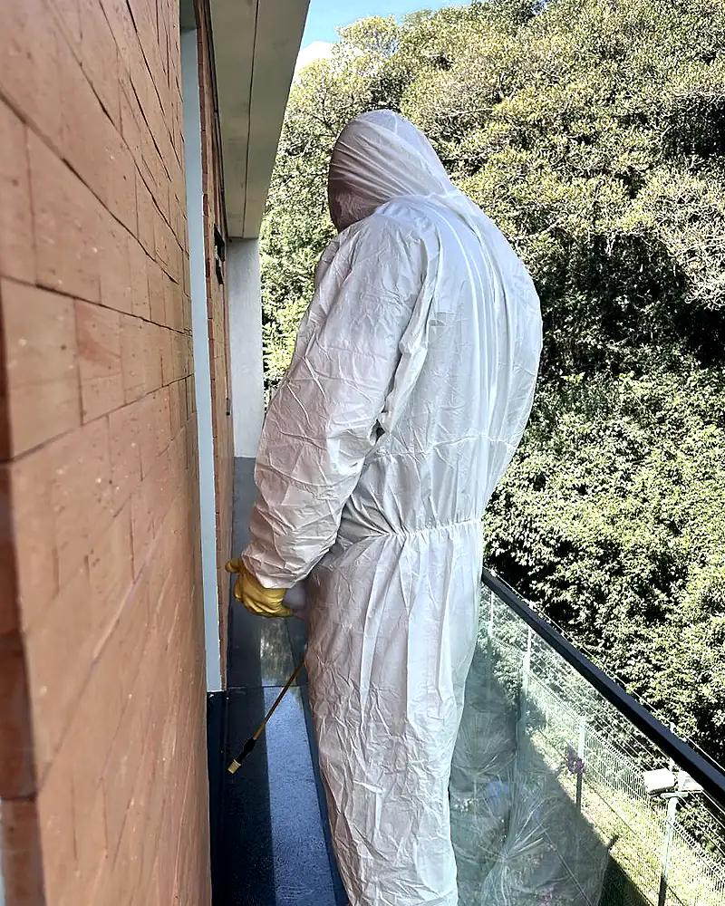 Técnico de macacão branco aplica produto ao longo do rodapé externo de uma sacada, com mata ao fundo.